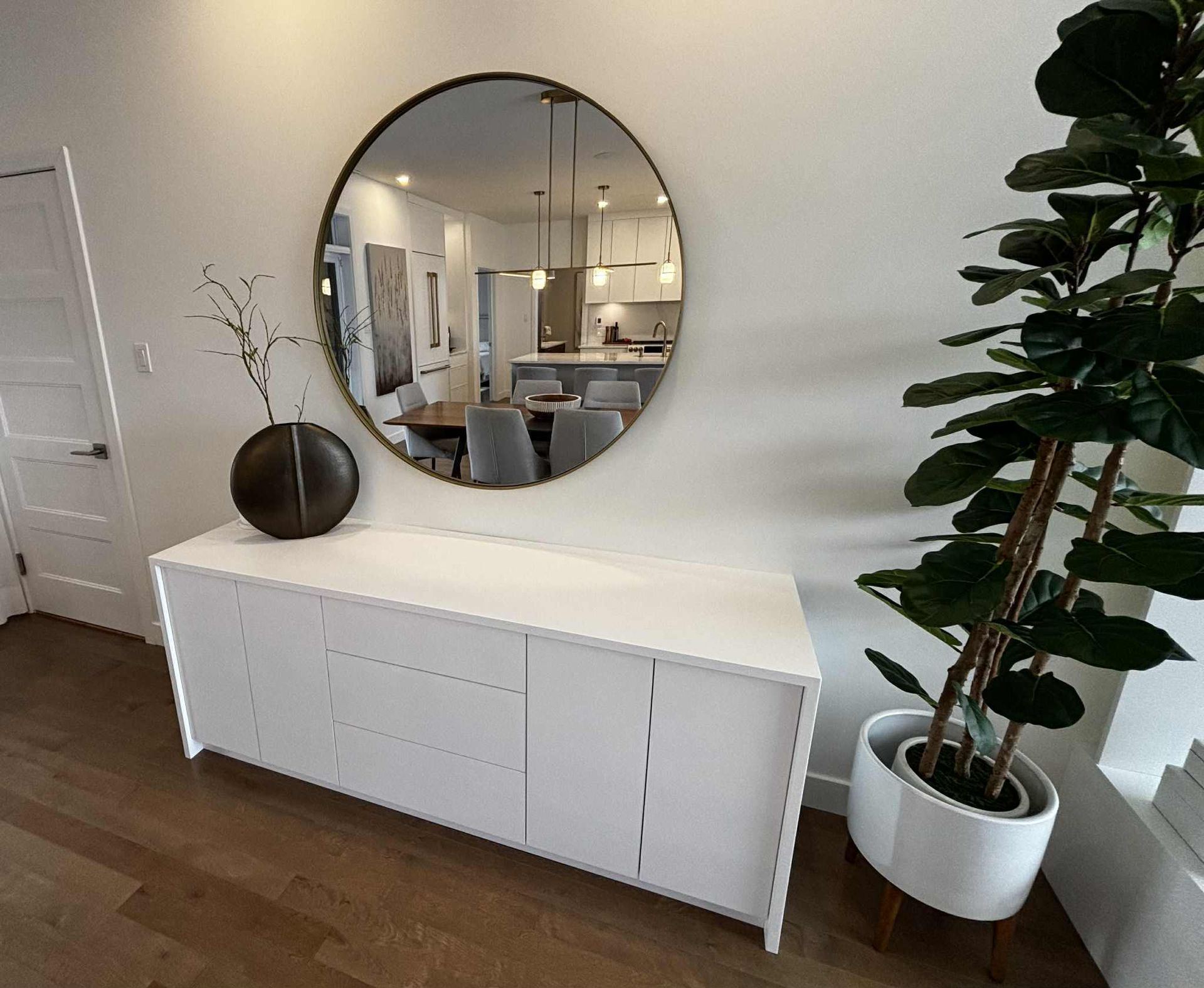 Buffet blanc avec miroir rond, plante et vase dans un intérieur bien éclairé.