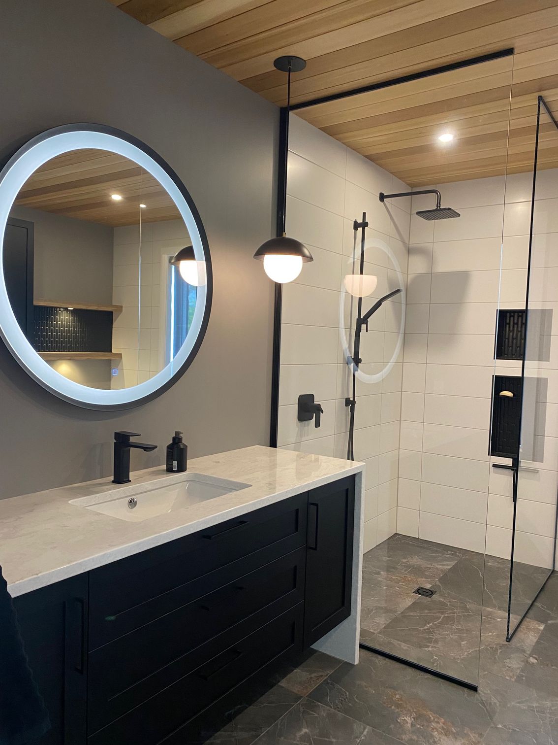 Salle de bains moderne avec meuble-lavabo noir, miroir rond, douche en verre et plafond en bois.