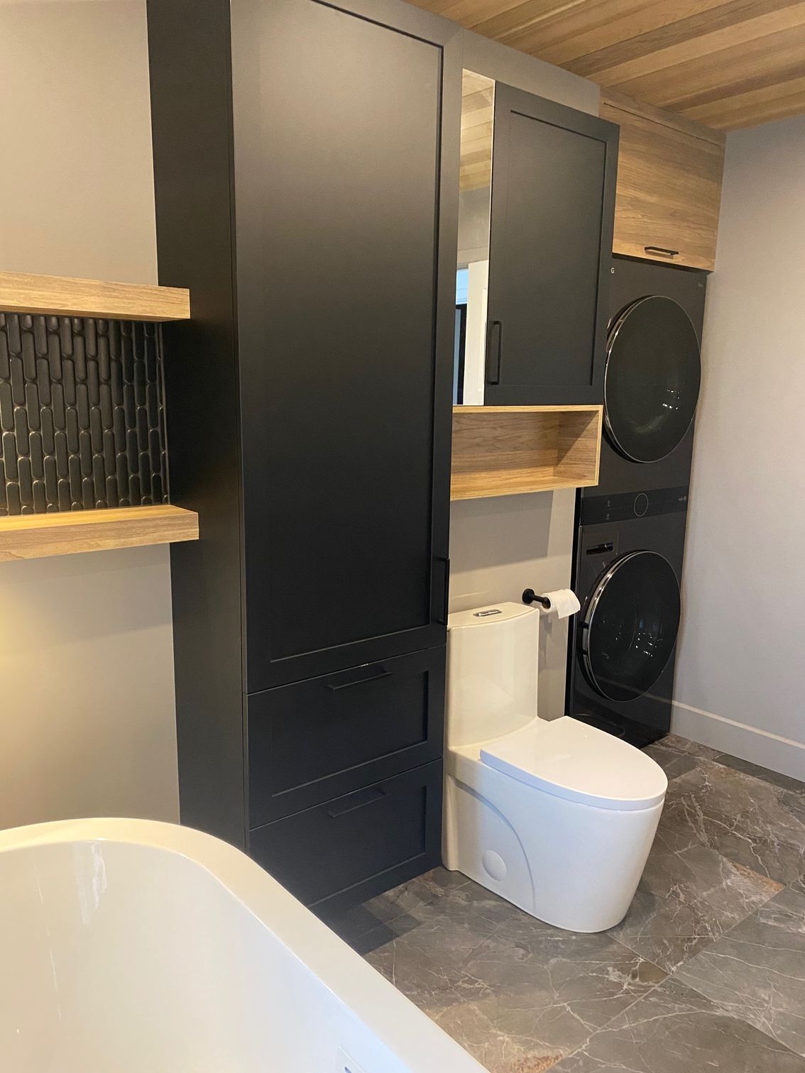 Salle de bains moderne avec meubles noirs, toilettes, lave-linge/sèche-linge superposés et baignoire.