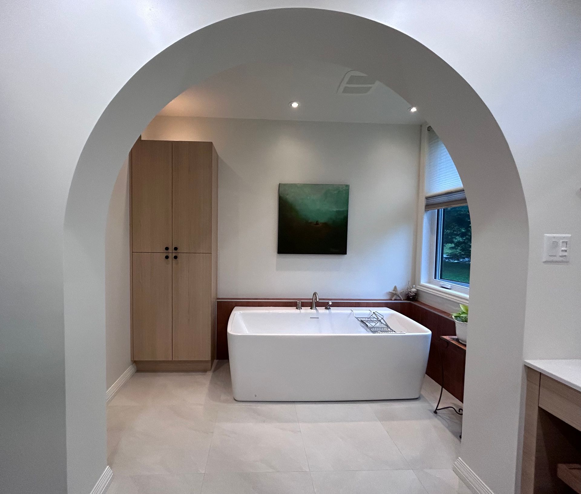 Salle de bains avec arche, murs blancs, baignoire îlot, meuble en bois et fenêtre.