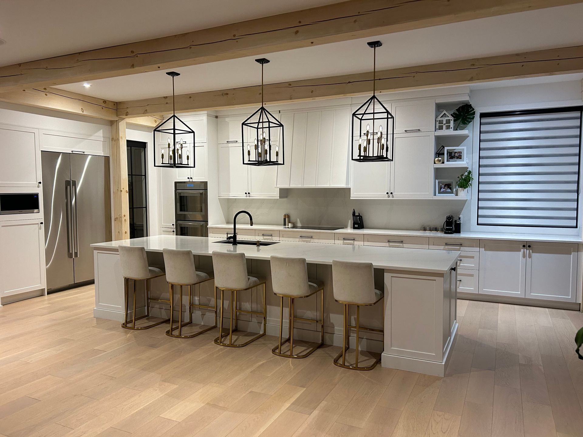 Cuisine blanche moderne avec îlot central, luminaires suspendus, appareils électroménagers en acier inoxydable et poutres apparentes.