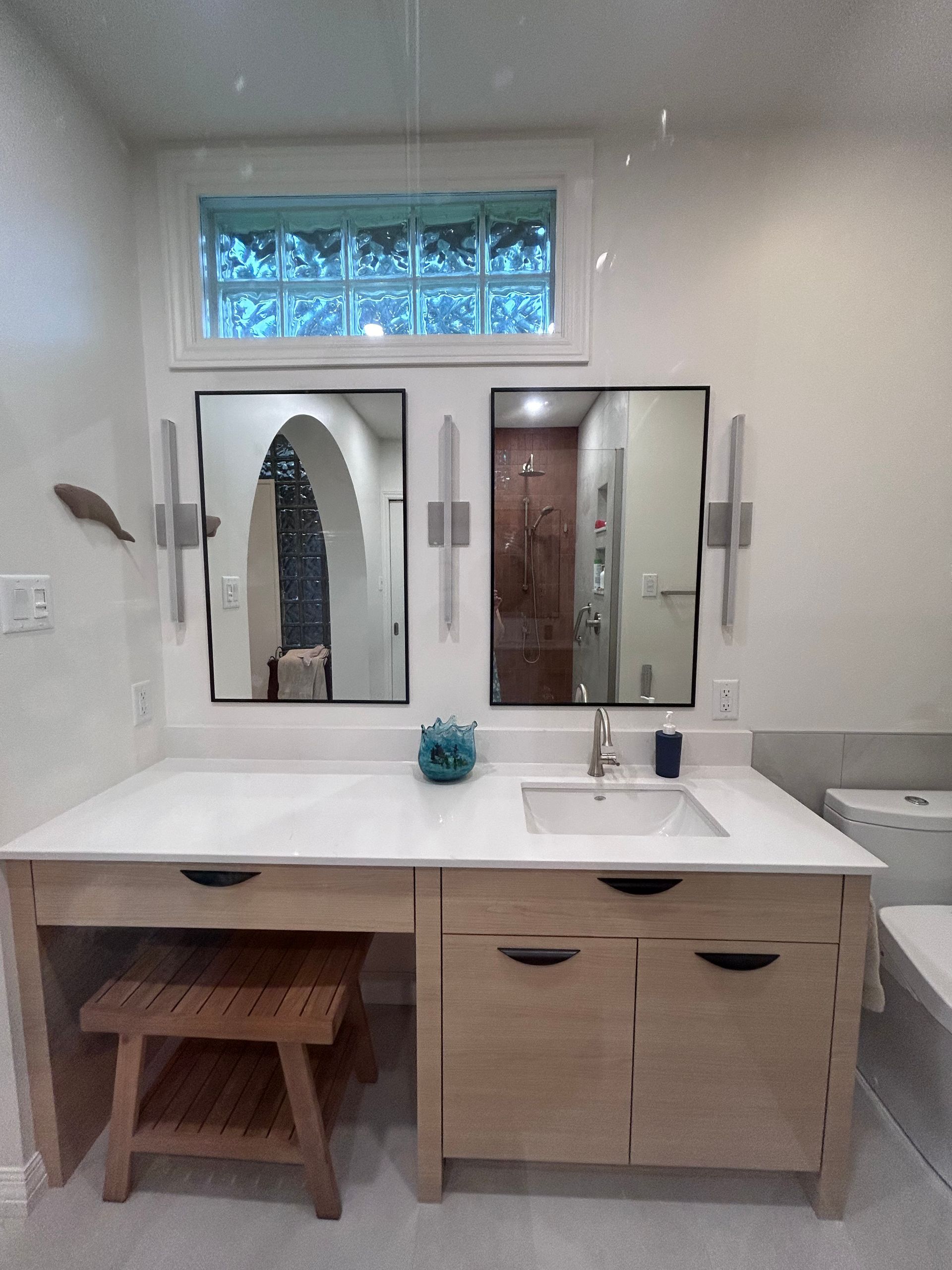 Meuble-lavabo de salle de bain avec armoires en bois clair, deux miroirs et fenêtre en pavés de verre bleu.