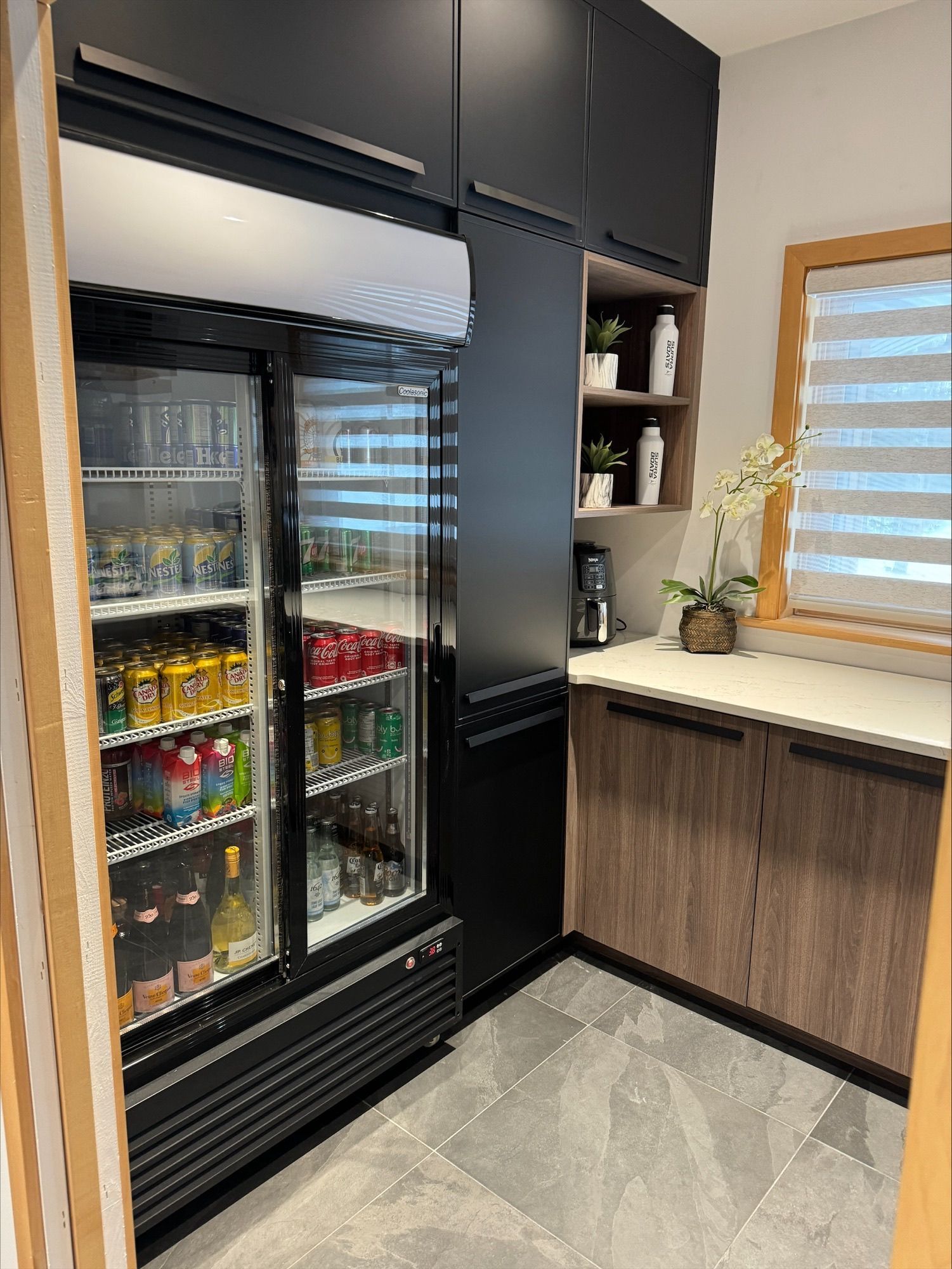 Garde-manger avec un réfrigérateur commercial rempli de boissons, des armoires noires et des éléments en bois.
