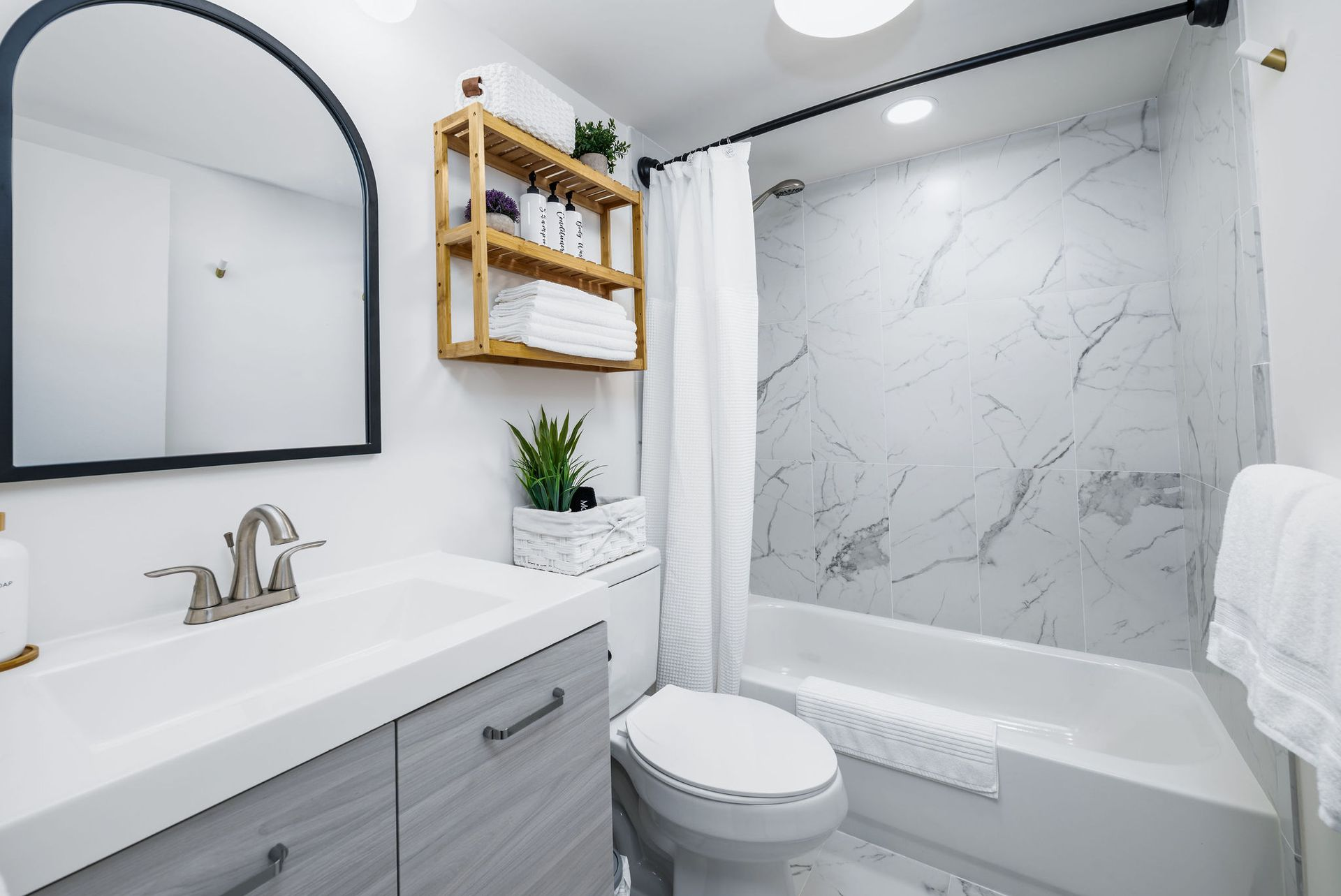 Bathroom with white walls, marble shower, wooden shelf with toiletries, arched mirror, and gray vanity.