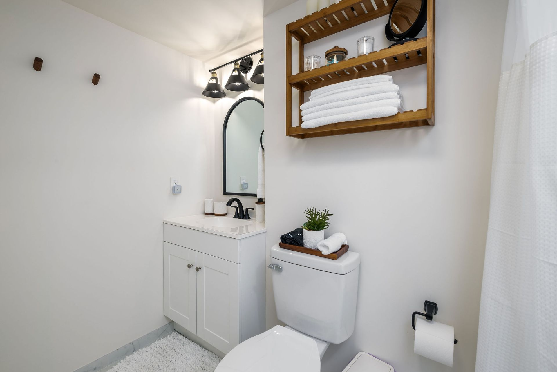 Small white bathroom with toilet, vanity, and wooden shelves holding towels and decor.