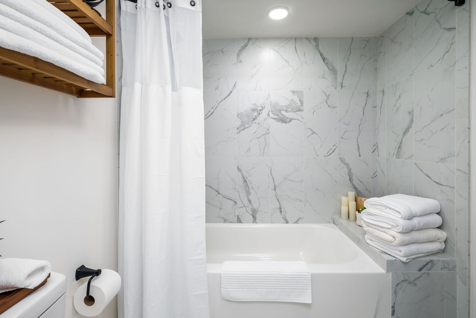 Bathroom with a white bathtub, marble tile, white shower curtain, and stacked towels.