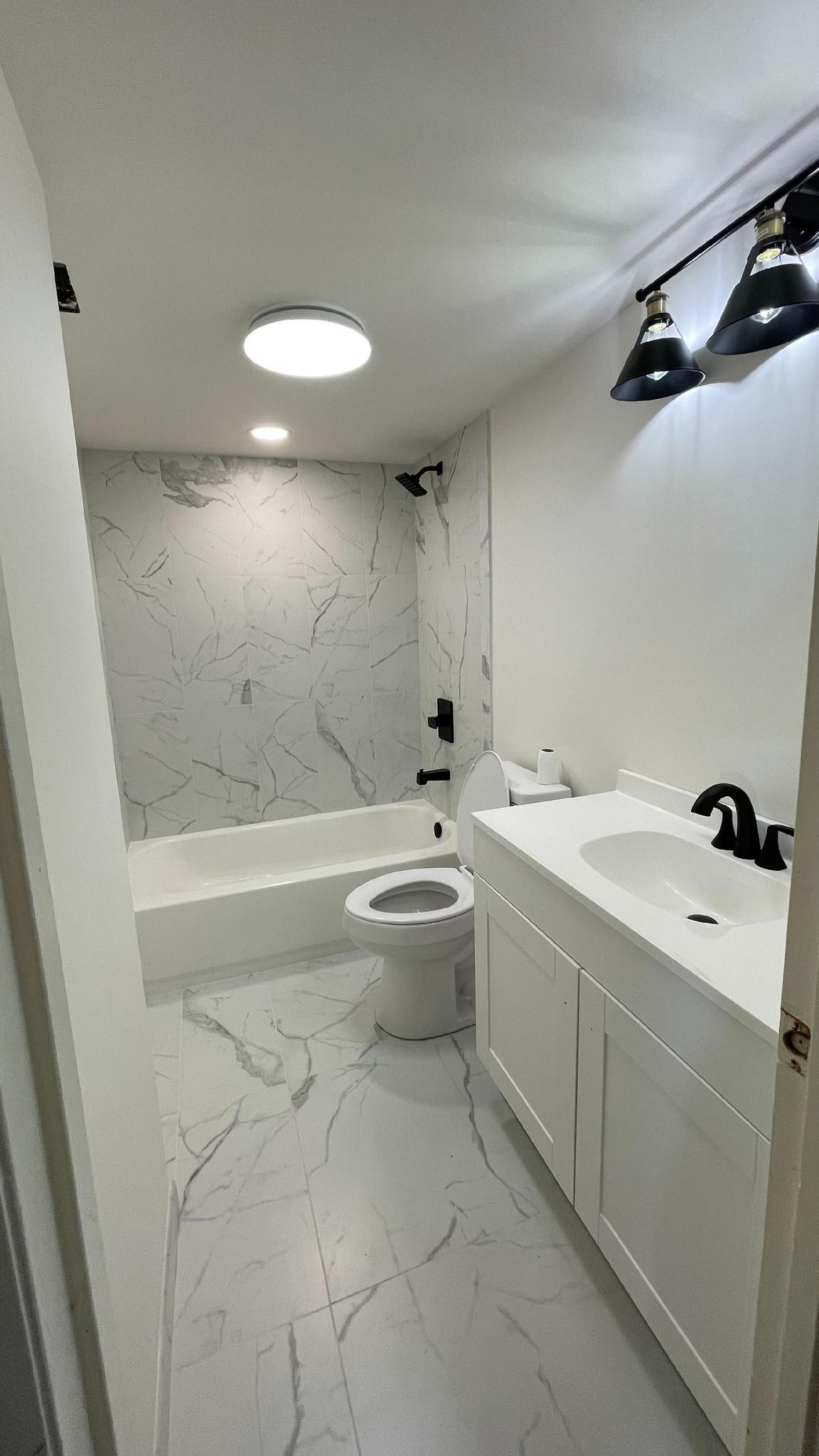 White bathroom with marble-look tile, tub, toilet, vanity, and black fixtures; overhead lighting.
