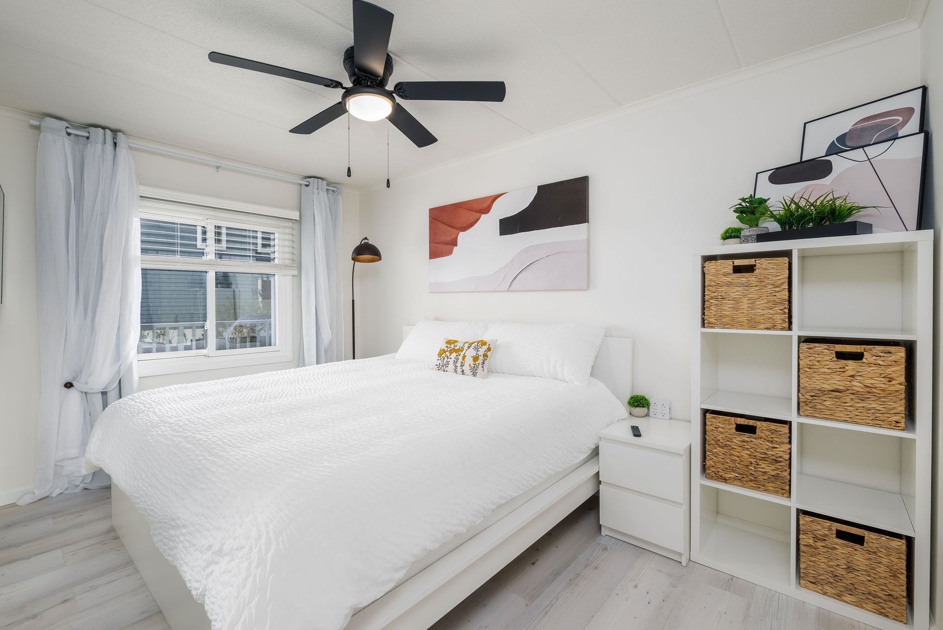 Bright bedroom with a bed, dresser, shelves, ceiling fan, and abstract artwork.