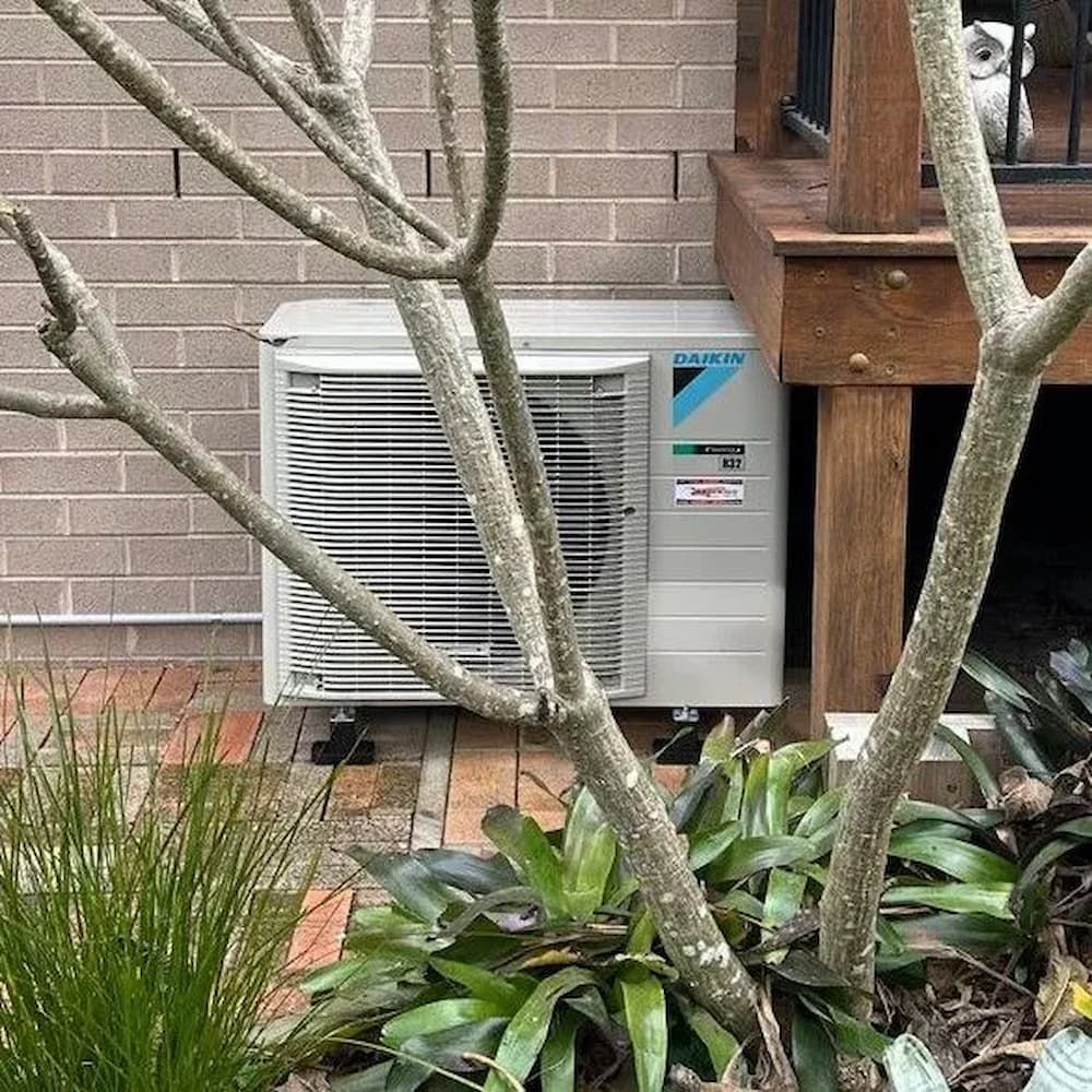 A Daikin Air Conditioner Is Sitting On The Side Of A Brick Building Next To A Tree — Mackies In Taree, NSW