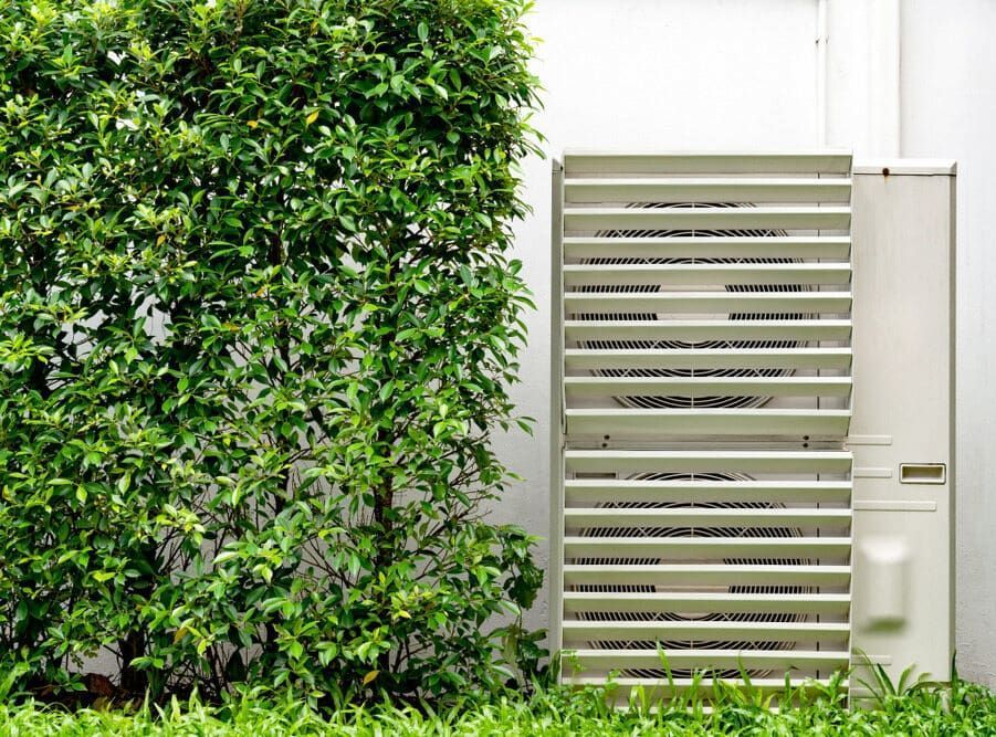 A White Air Conditioner Is Sitting On The Side Of A Building Next To A Bush — Mackies In Taree, NSW