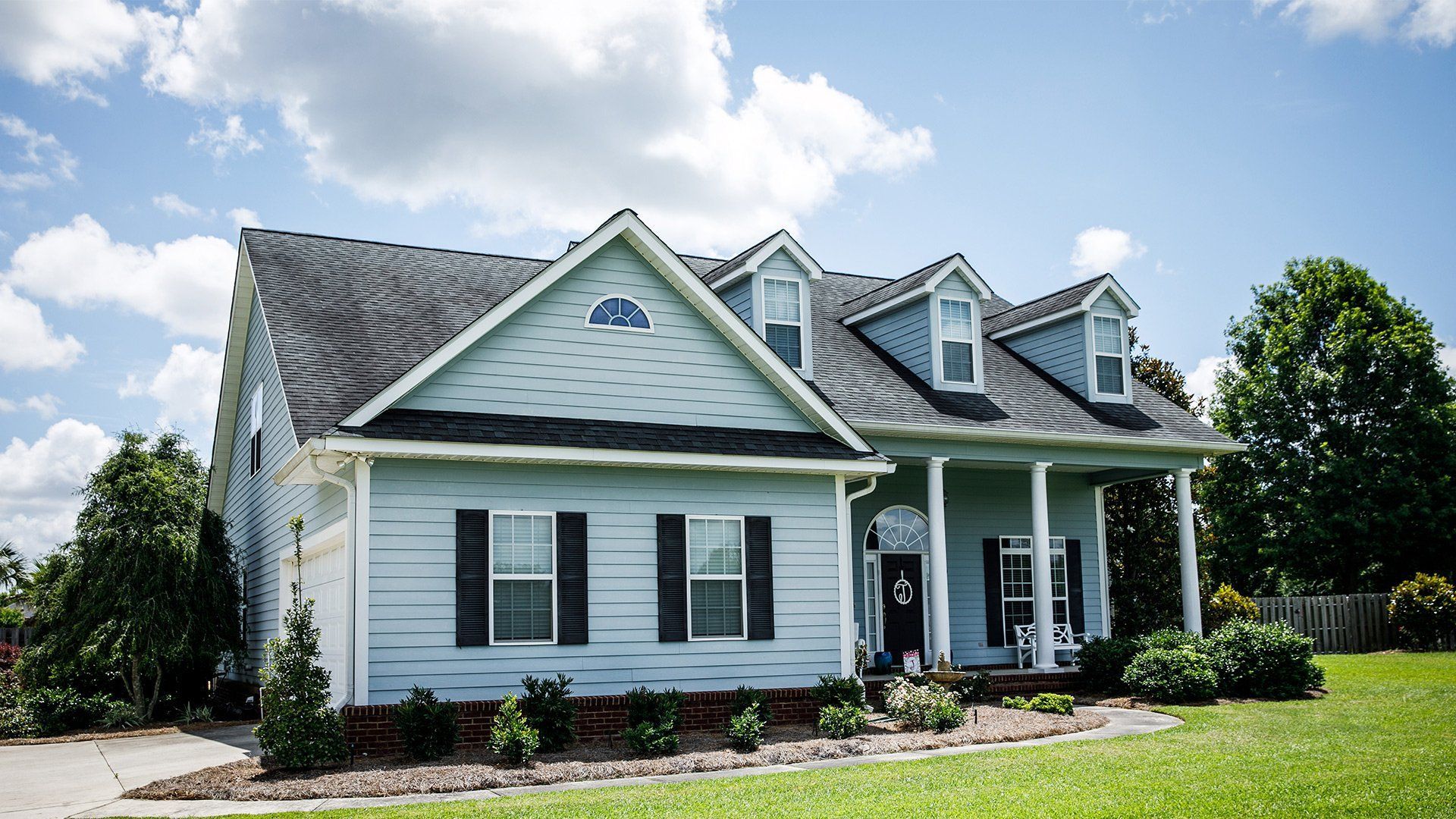 White suburban house with dark shutters, front porch, and landscaped lawn under a blue sky