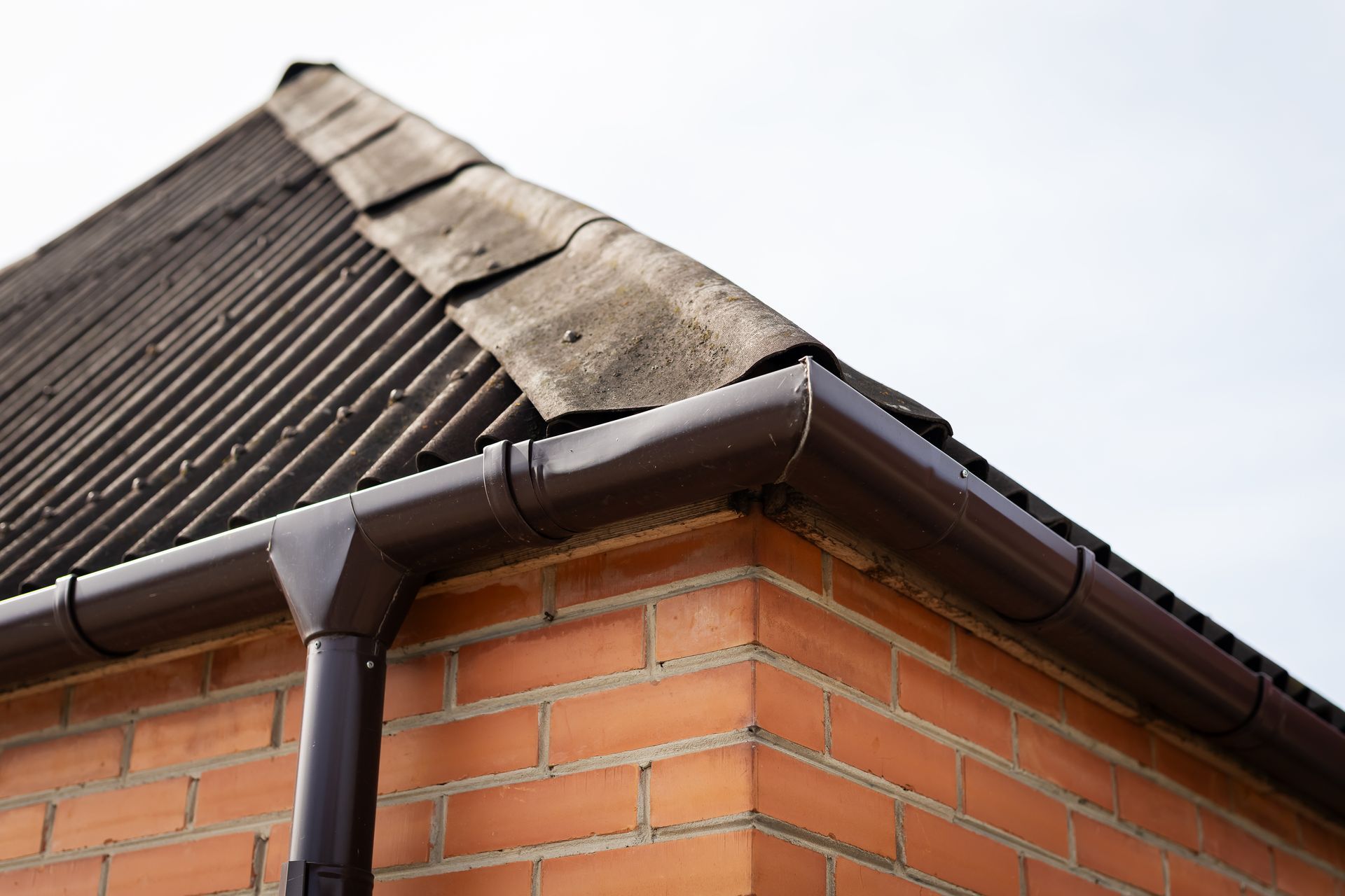 Corner of a brick house with dark roof tiles and a rain gutter under a cloudy sky Corner of a brick house with dark roof tiles and a rain gutter under a cloudy sky