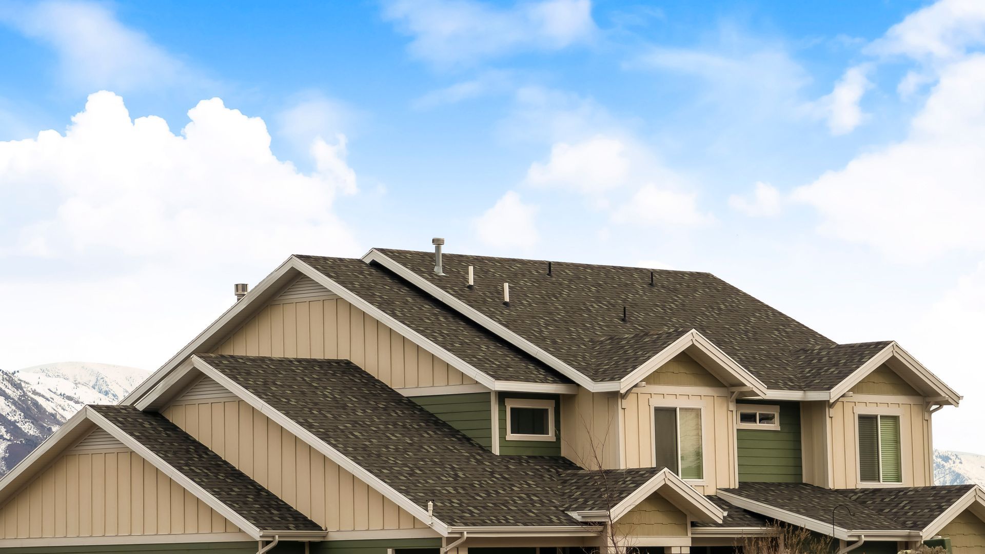 Suburban house roof with beige siding and dark shingles under a blue sky with clouds