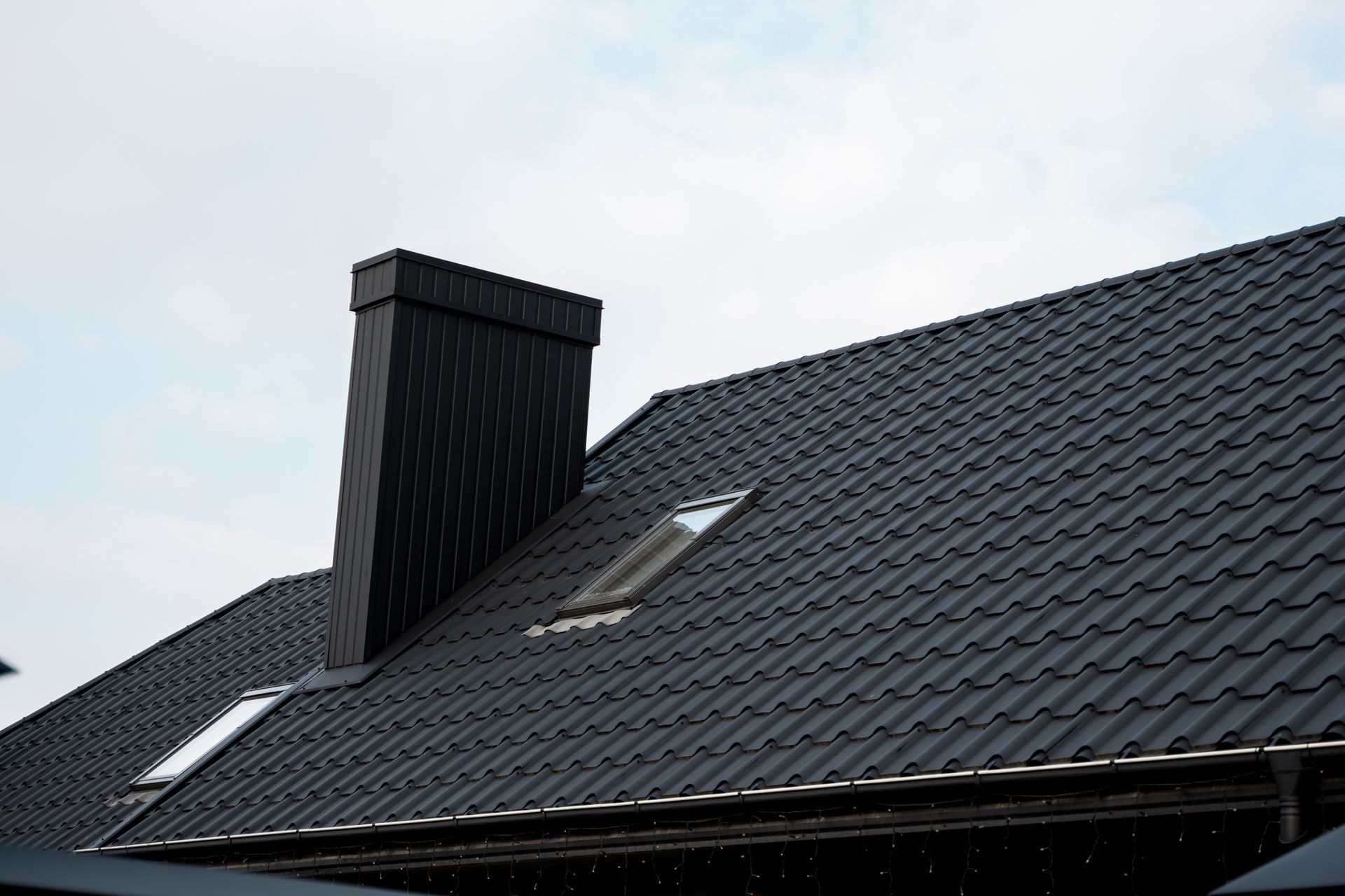Dark gray tiled roof with a black chimney and skylight against a cloudy sky