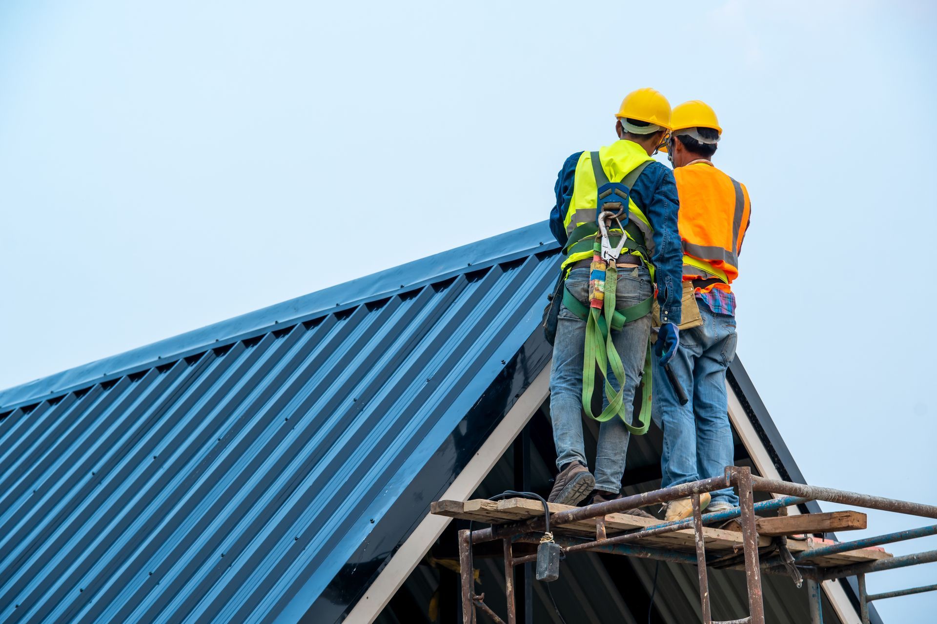 Two workers on a scaffold installing a blue metal roof, wearing safety harnesses and hard hats