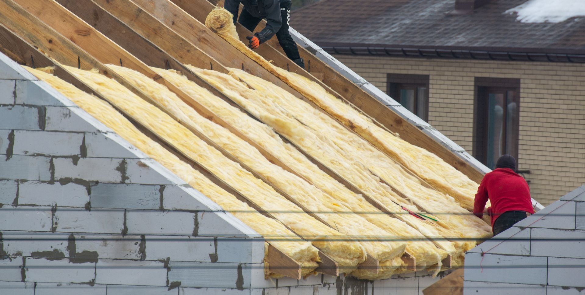 Worker installing insulation on a sloped roof beside a brick house under construction Worker installing insulation on a sloped roof beside a brick house under construction