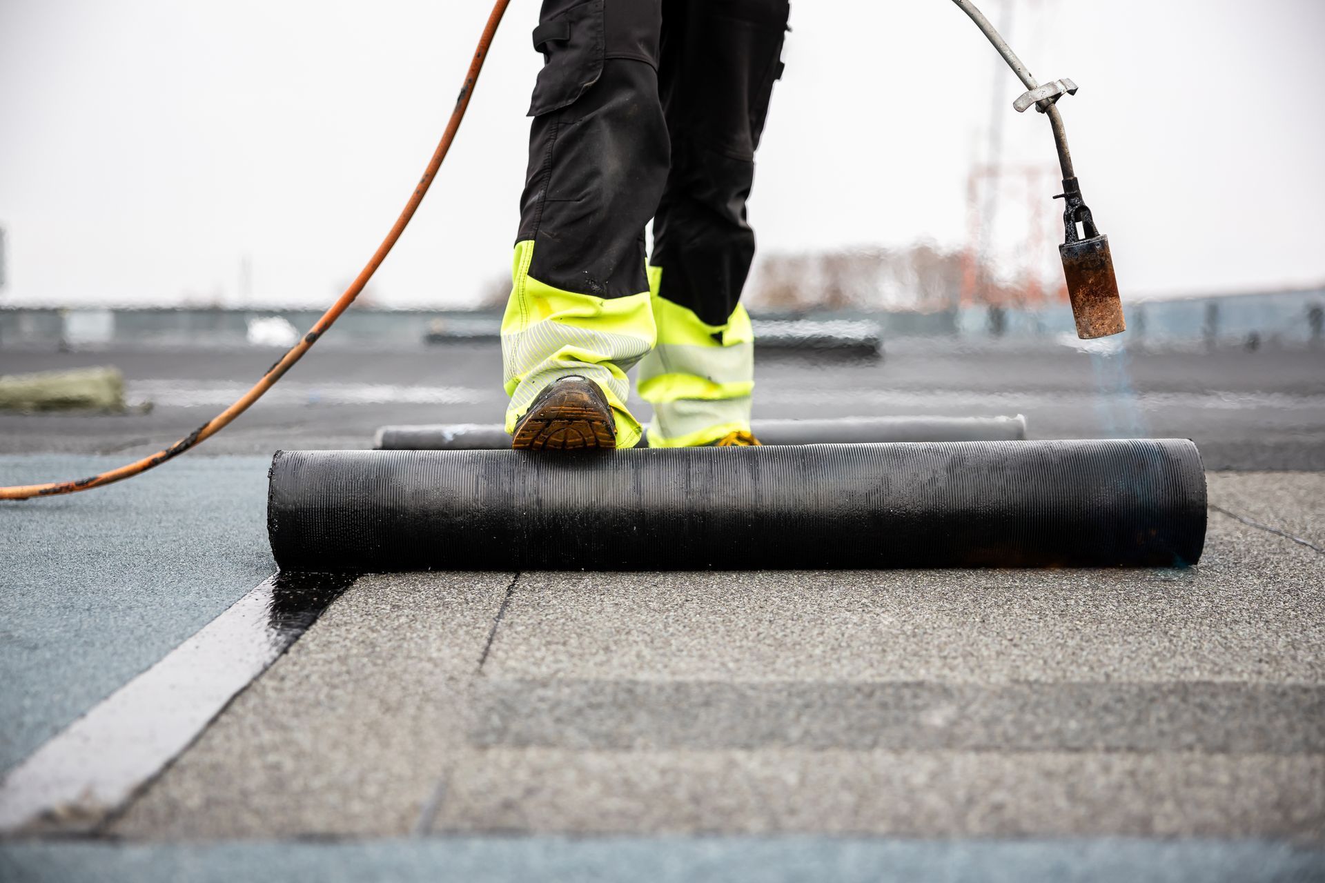 Worker installing black roofing membrane with a torch on a flat roof, wearing bright yellow boots