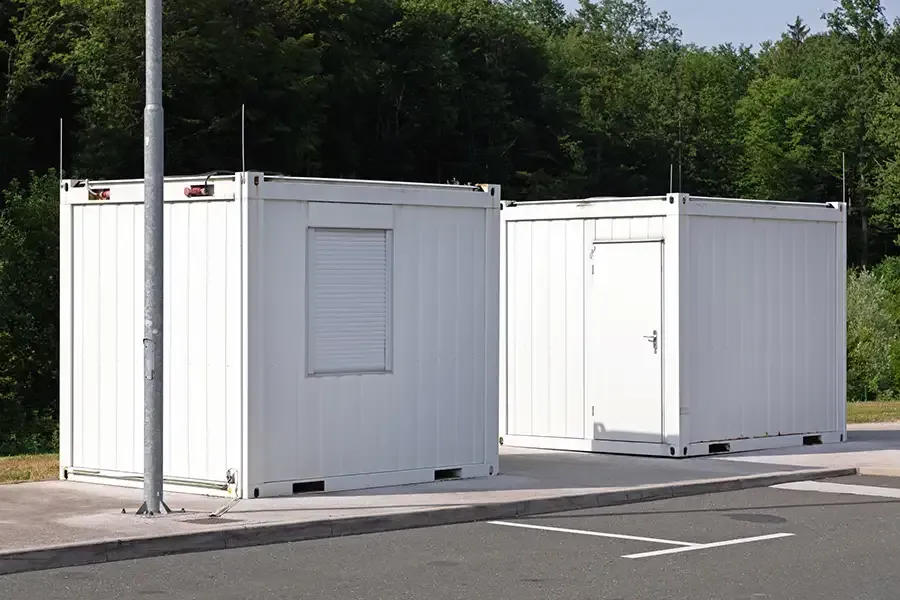 Two white shipping containers on pavement, one with a window, the other with a door. Trees in the background.