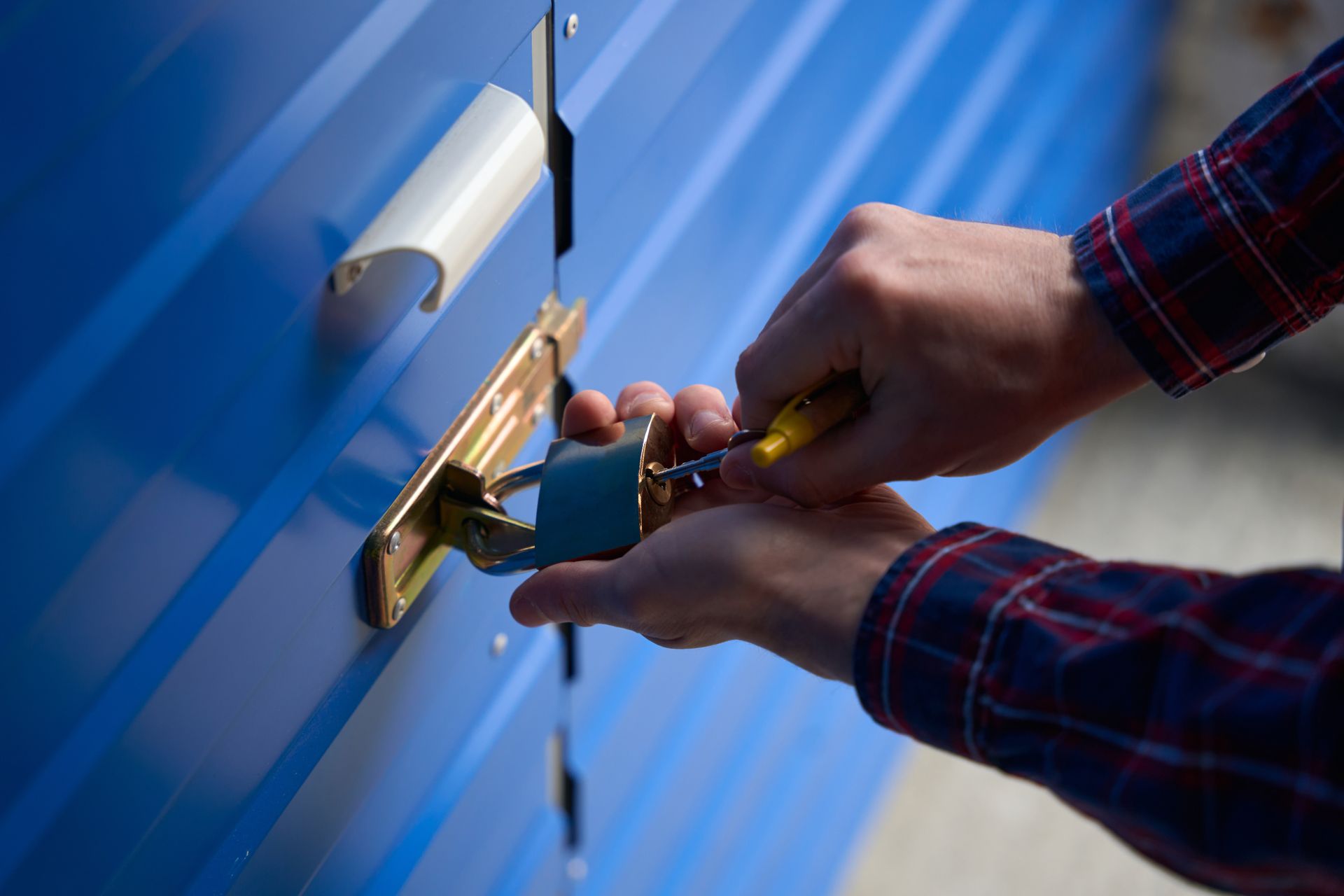 Hands using a screwdriver to unlock a padlock on a blue storage unit door.