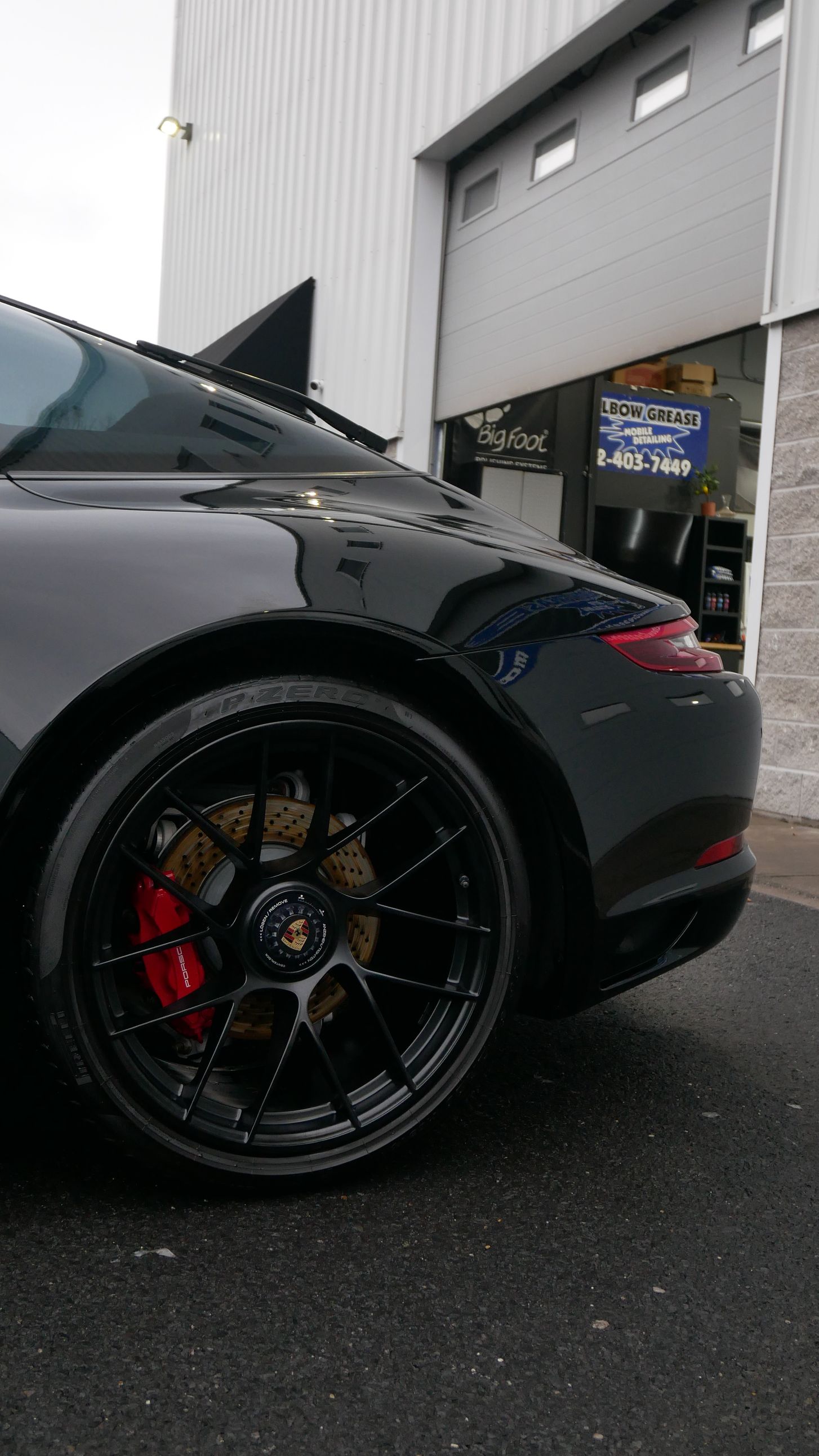 Black sports car with custom black wheels and red brake calipers parked by an industrial garage.