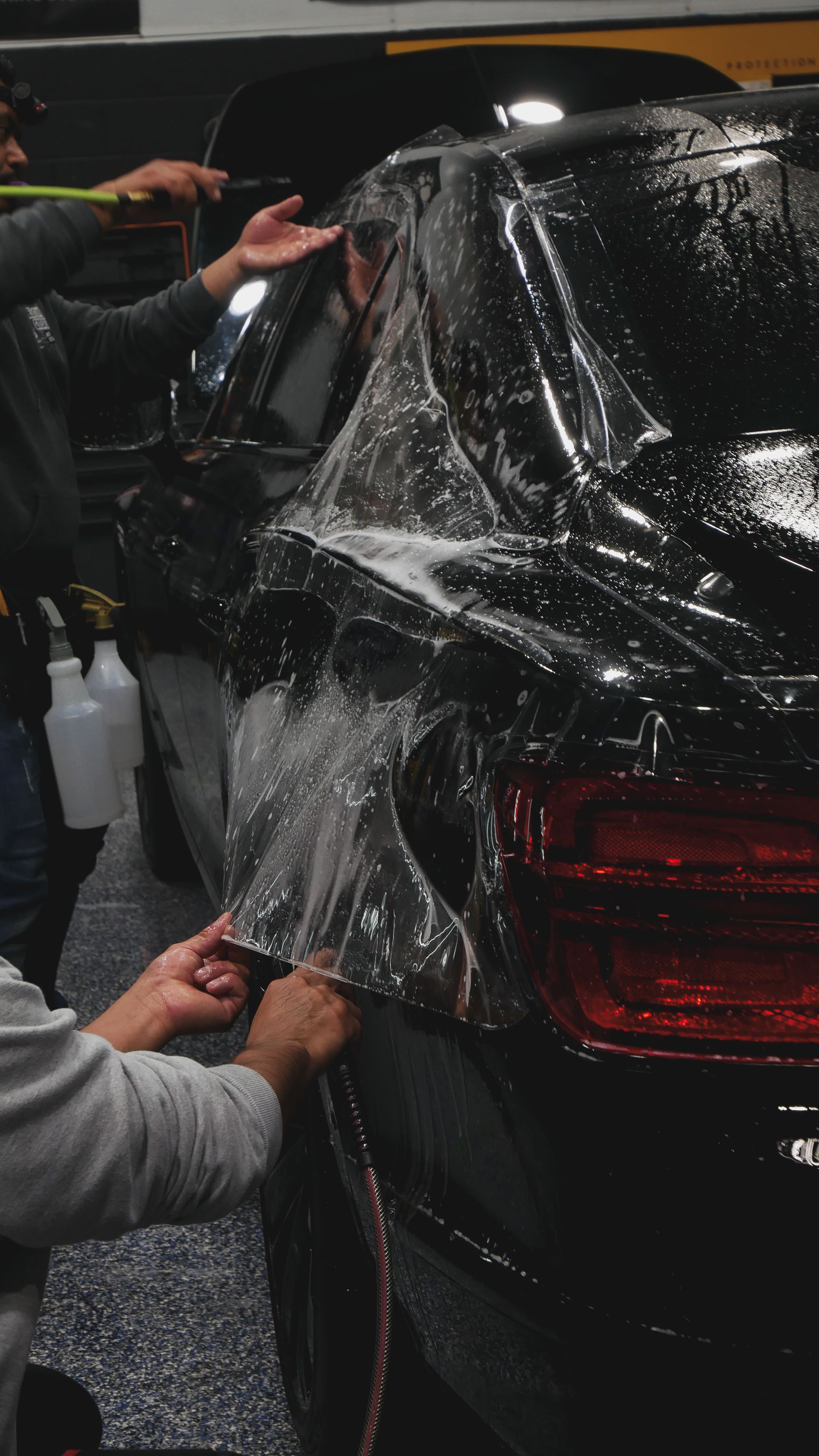 Two people applying clear film to the rear side of a black SUV indoors