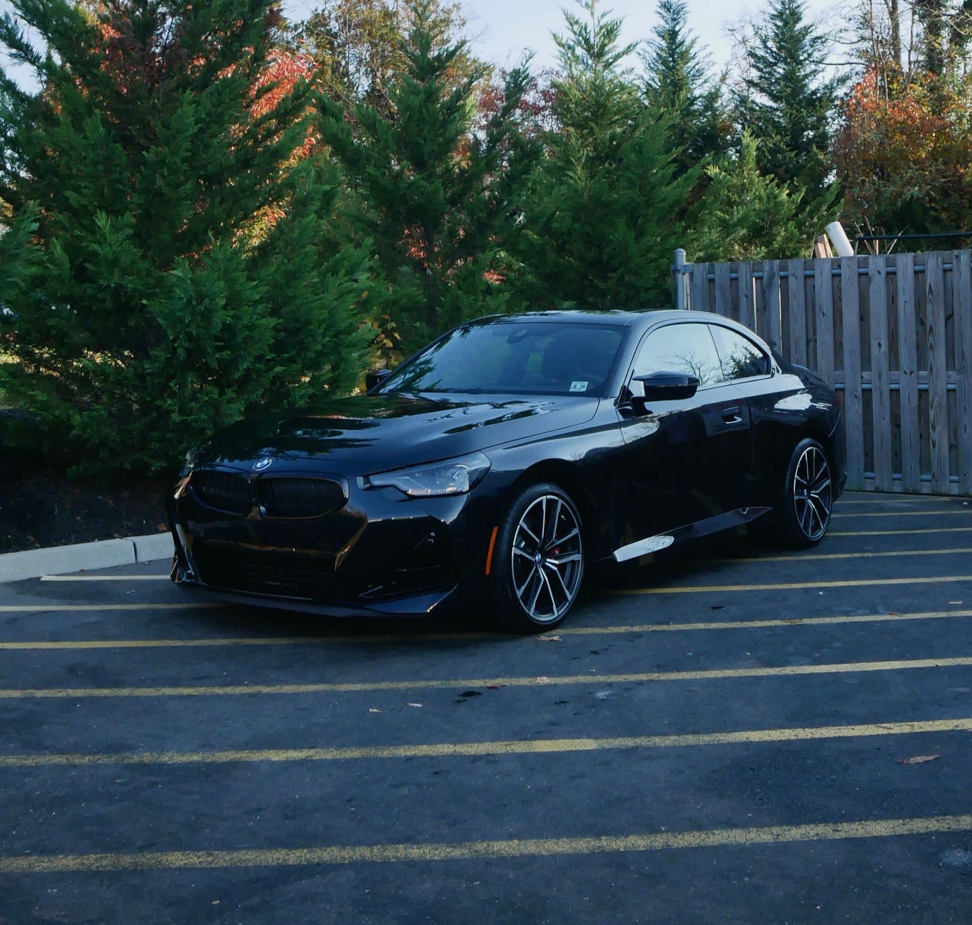 Black sedan parked in a lot beside a wooden fence, with evergreen trees in the background.