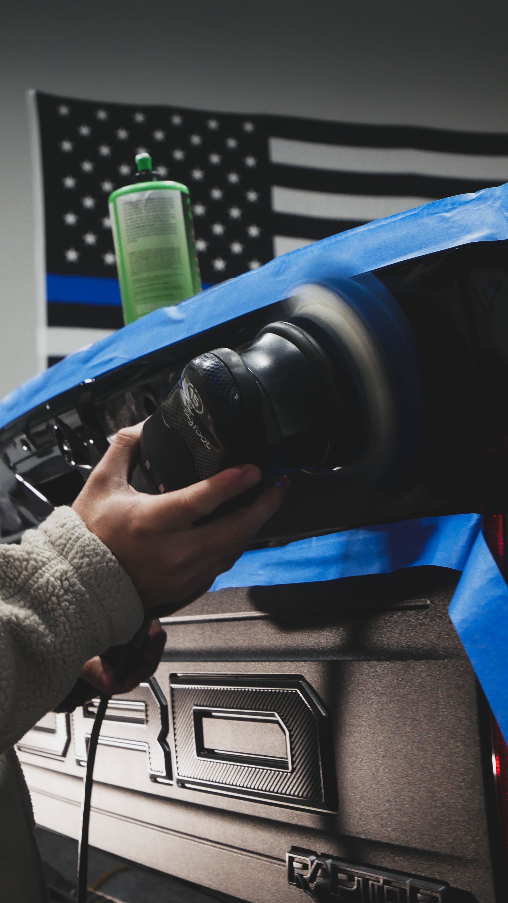 Hand holding a black car part over a blue-taped workbench with an American flag in the background