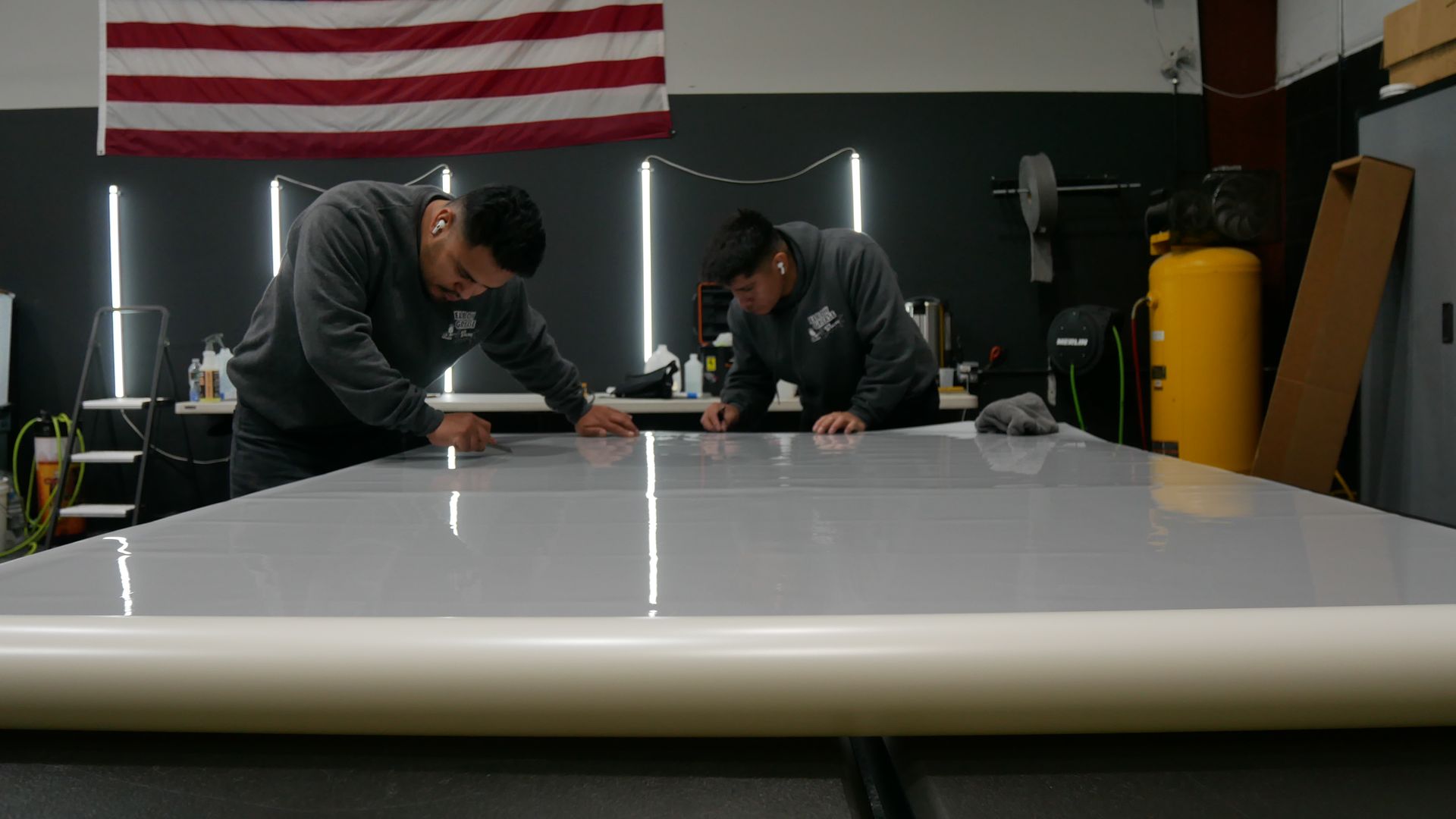 Two workers measuring a large white panel in a workshop with an American flag in the background