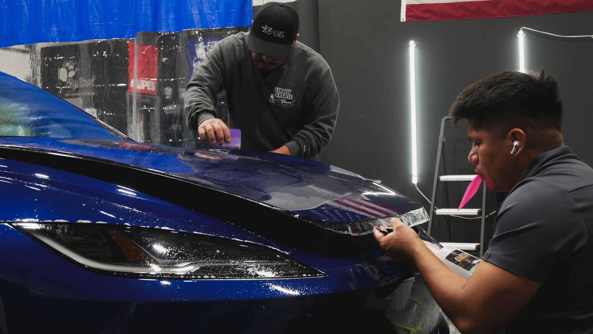 Two workers apply glossy wrap to a blue sports car in a workshop