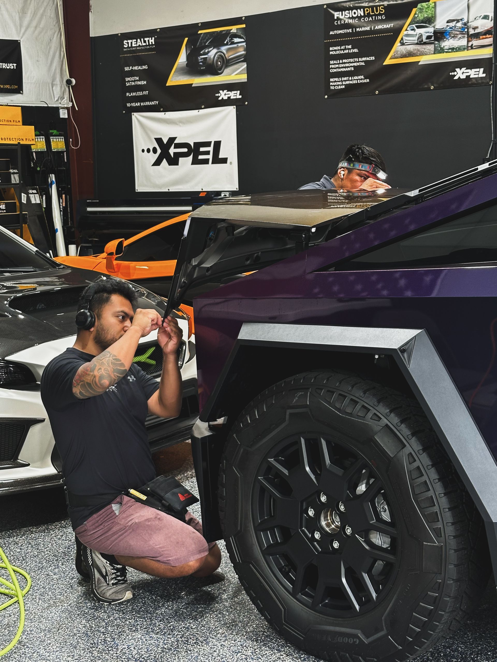 Mechanic kneels beside a purple off-road vehicle, working under the front fender in a garage.