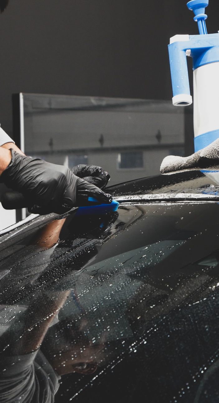 Gloved hand washing a black car with blue foam and water spray in a car wash.