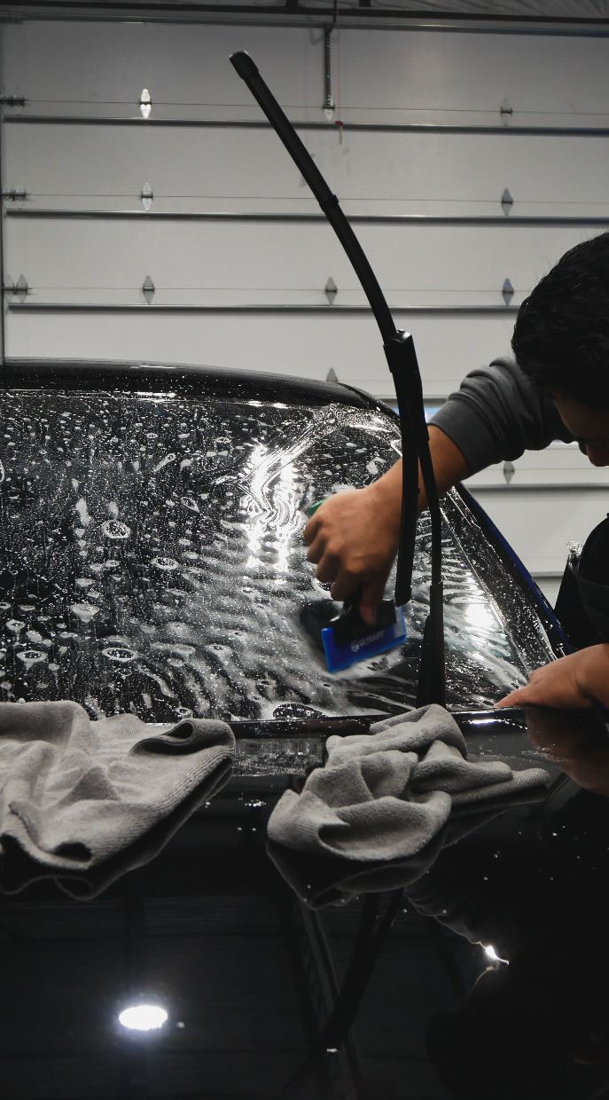 Person pressure-washing a car windshield in a garage, with water spray and towels on the hood