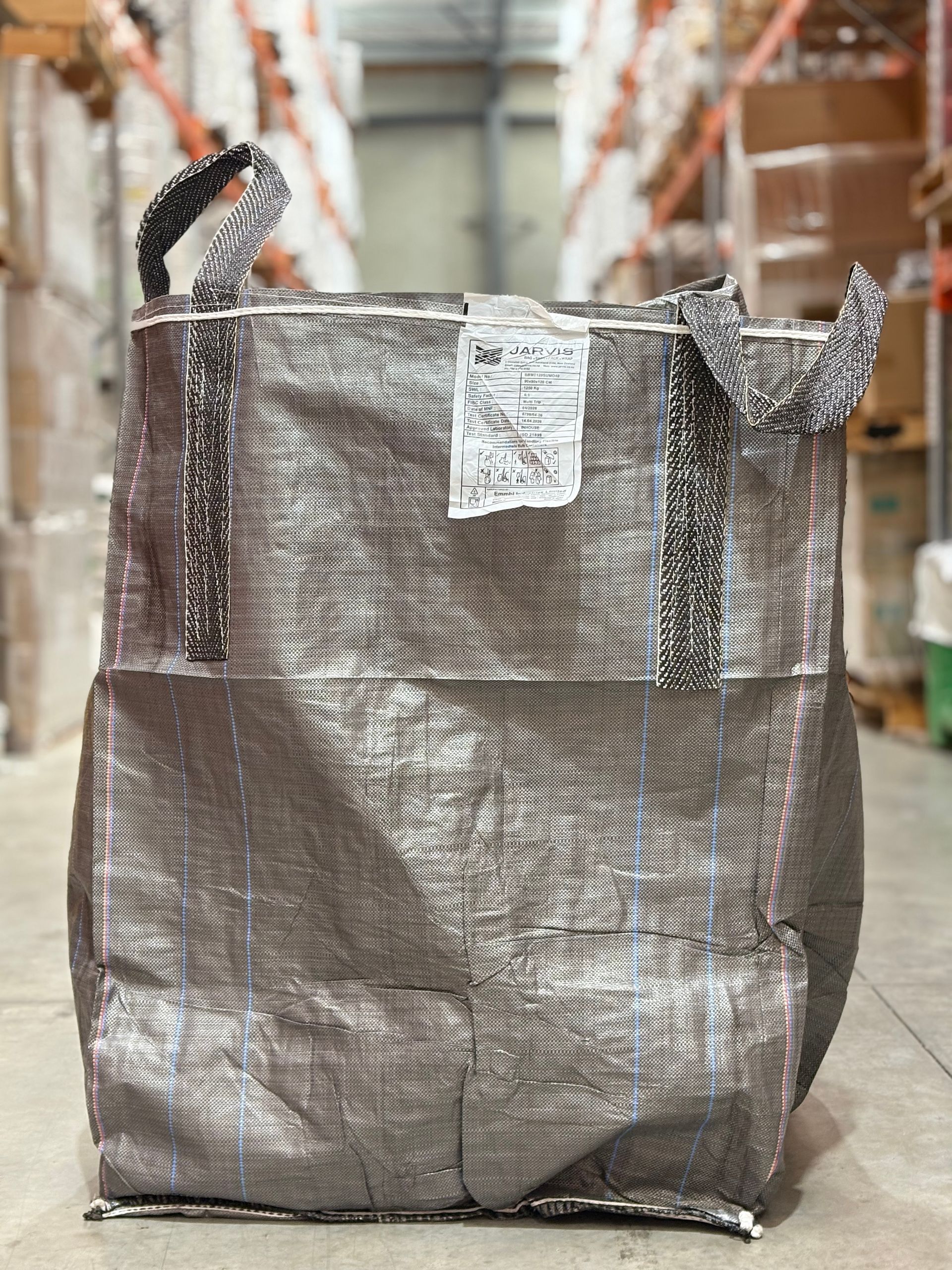 Large woven shopping bag on a warehouse floor aisle, with handles and a shipping tag attached.