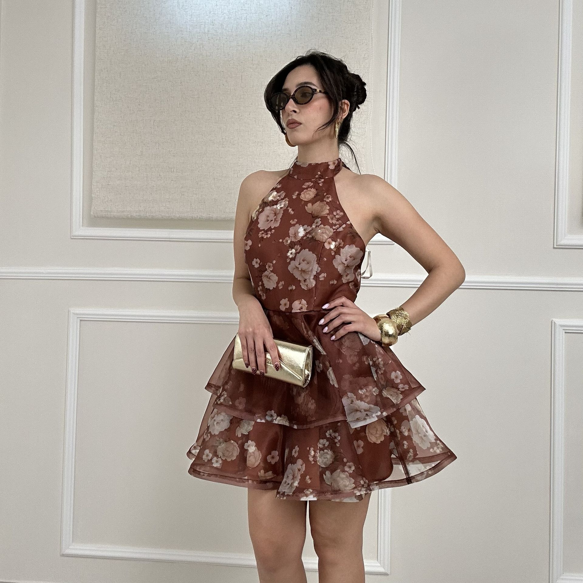 Woman in a brown floral dress and sunglasses poses against a paneled wall, holding a gold clutch.