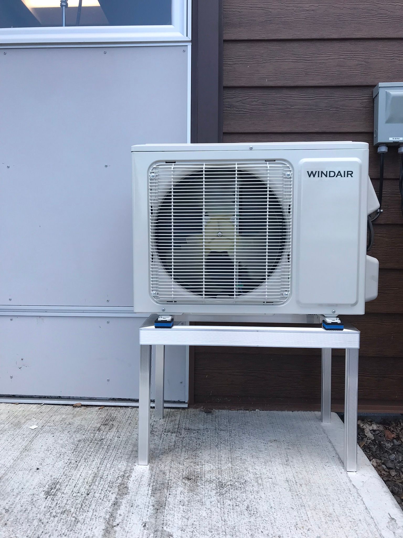 Unité de climatisation blanche sur un support métallique, fixée près du mur d'un bâtiment.