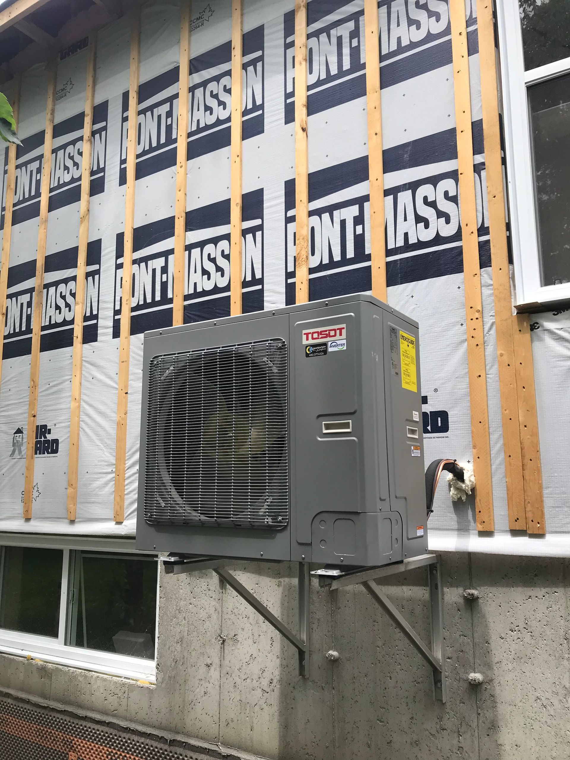 Extérieur d'un bâtiment avec une unité de climatisation grise fixée sur un mur en béton. L'unité est en cours d'installation.
