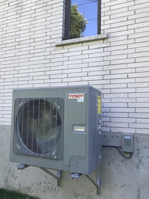 Unité de climatisation grise fixée sur un mur de briques sous une fenêtre.