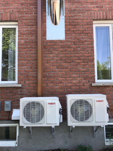 Deux unités de climatisation blanches sont fixées sur un mur de briques, sous les fenêtres. Une cheminée s'élève au-dessus.