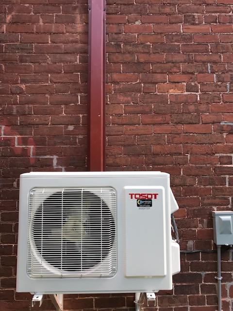 Climatiseur Tosot blanc sur un mur de briques rouges, avec un conduit métallique rouge.