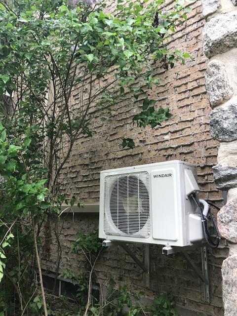 Unité de climatisation blanche fixée sur un mur extérieur texturé, avec un arbre à gauche.