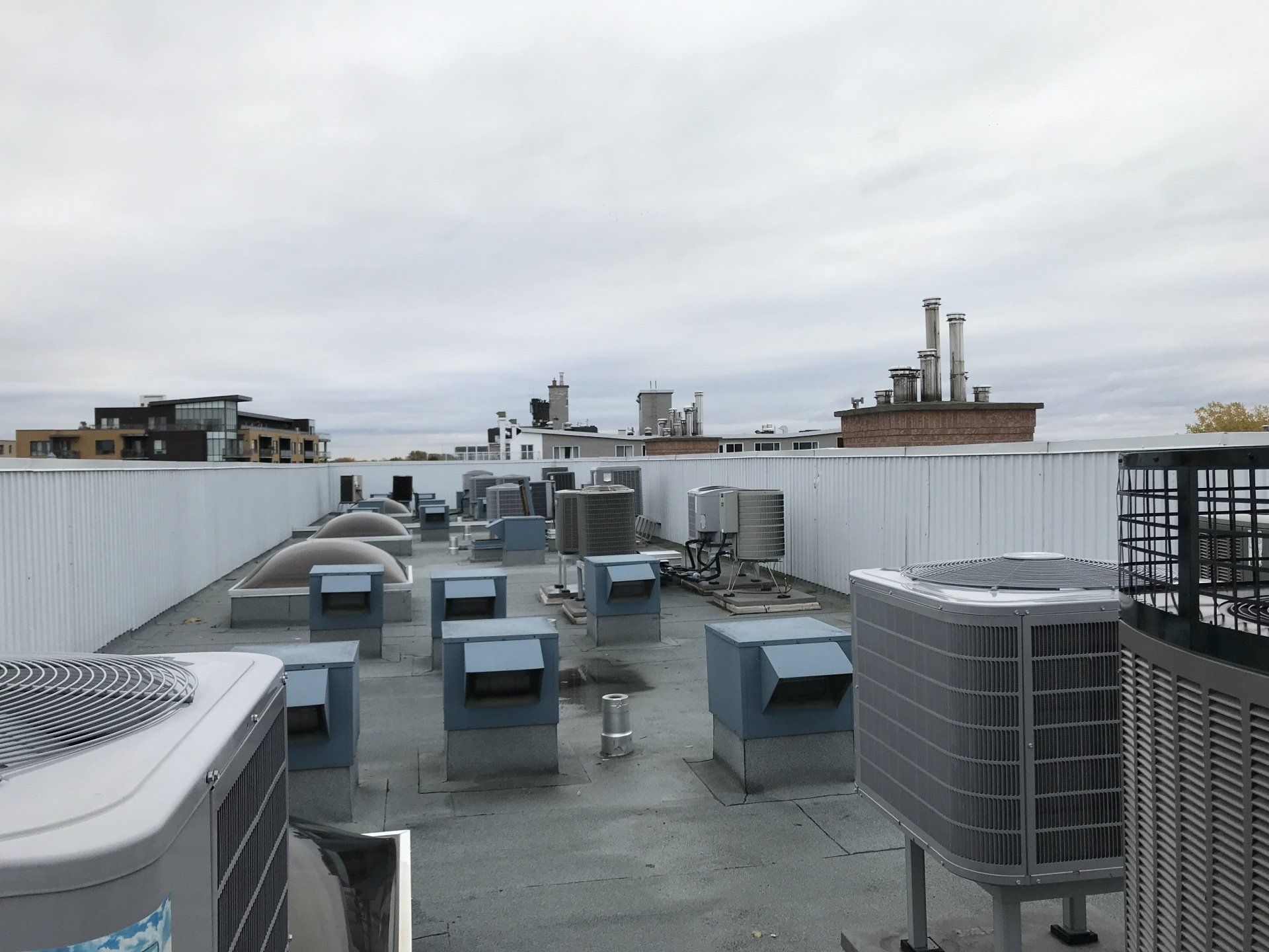 Toit-terrasse avec unités de climatisation, puits de lumière et muret blanc. Ciel couvert avec bâtiments en arrière-plan.