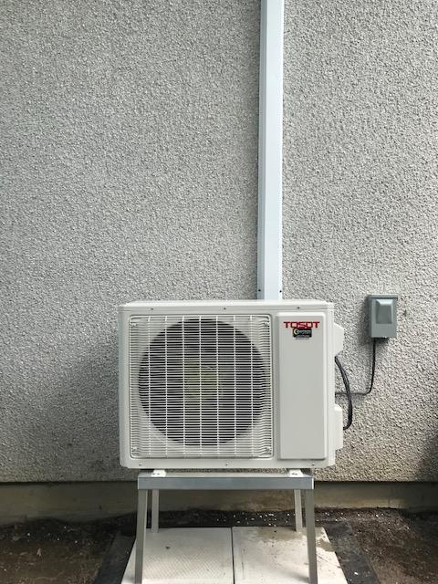 Unité de climatisation sur support métallique contre un mur gris texturé, à côté d'un tuyau blanc et d'un boîtier électrique.