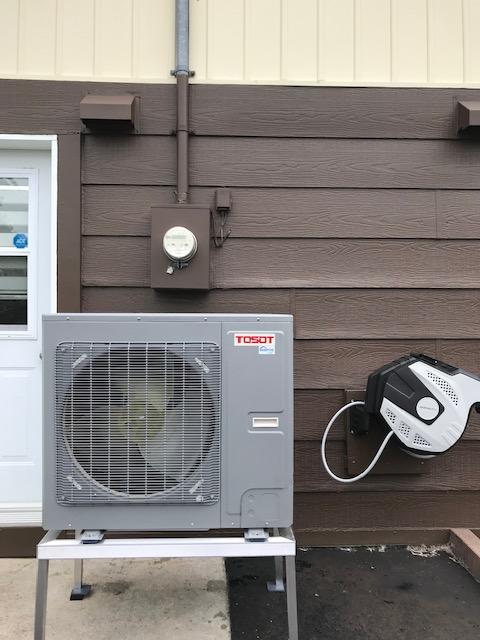 Unité de climatisation extérieure sur un support métallique, fixée sur un mur marron avec boîtier électrique et enrouleur de tuyau.