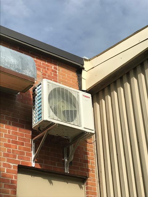 Un climatiseur extérieur monté sur un mur de briques, sous un toit.