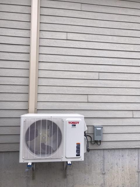 Unité de climatisation extérieure montée sur un mur de maison à bardage gris clair, avec conduit et boîte électrique.