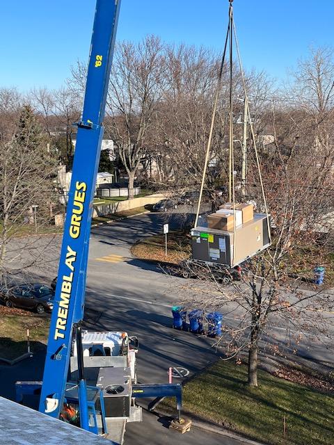 Une grue bleue soulève une unité de climatisation au-dessus d'une route bordée d'arbres et de maisons par une journée ensoleillée.