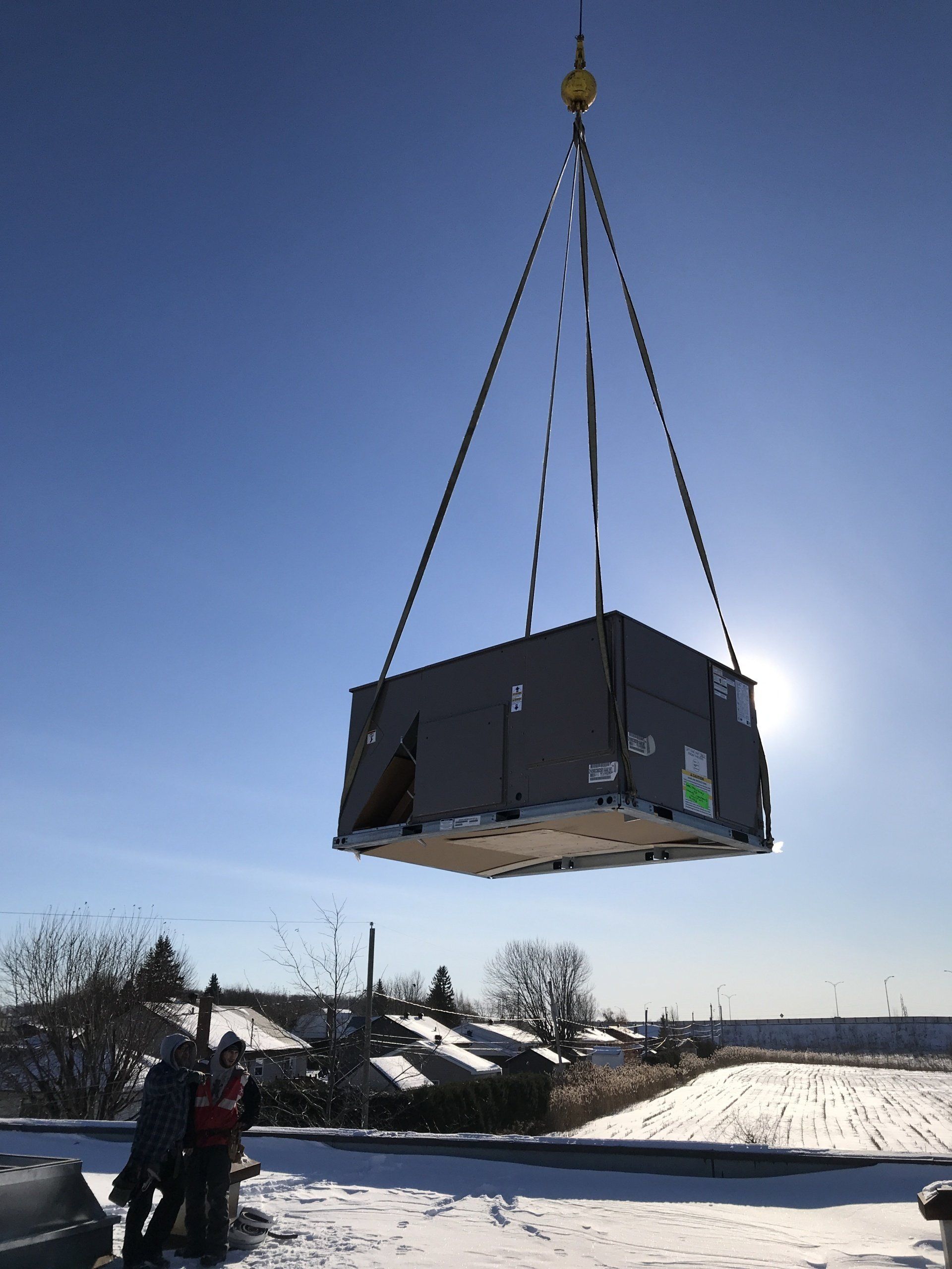 Par une journée ensoleillée, une grue soulève une grande boîte rectangulaire au-dessus d'un toit enneigé.