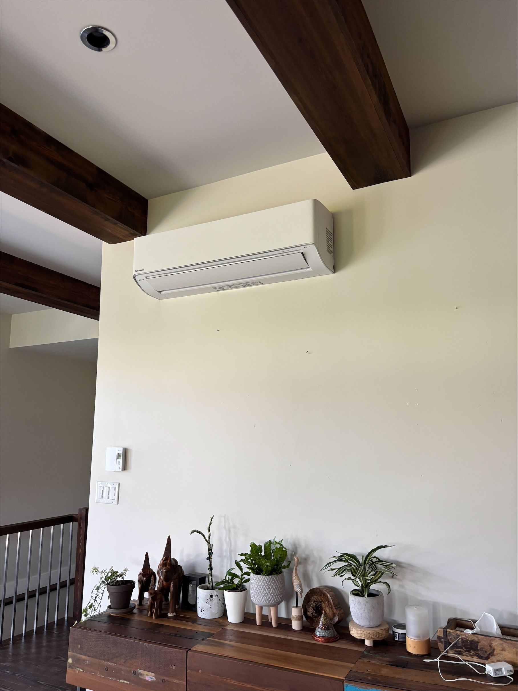 Climatiseur mural blanc au-dessus d'un meuble en bois avec des plantes. Poutres apparentes brunes au plafond.