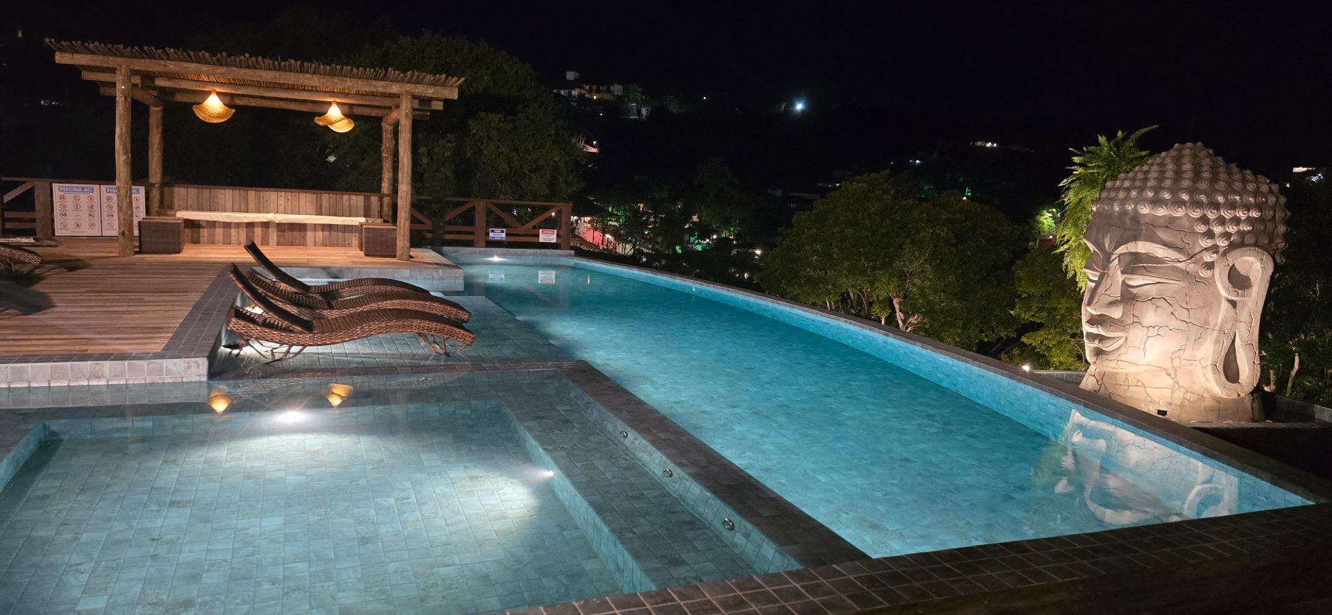 Piscina curva de resort em tons de turquesa, com deck de madeira, palmeiras e vista para as montanhas sob um céu azul