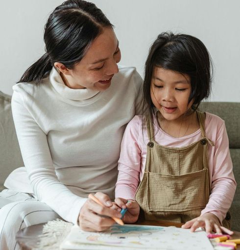 A woman is sitting next to a little girl who is drawing with colored pencils.